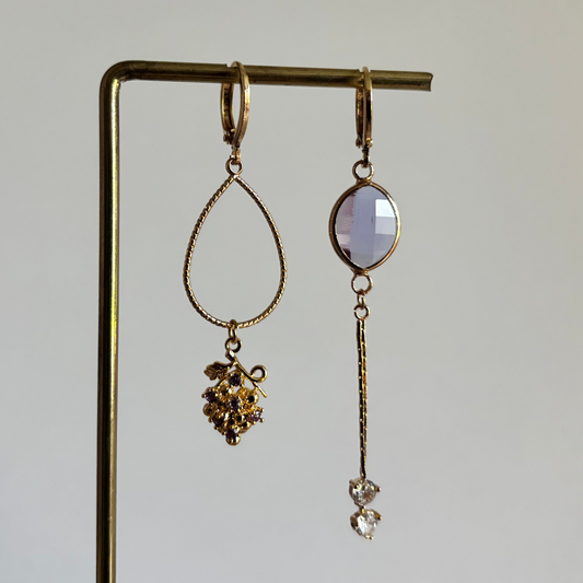 Set asymmetrische goudkleurige oorbellen aan een display. Links een ronde lila facetgeslepen steen met daaronder een fijn goudkleurig staafje en twee transparante steentjes. Rechts een grote opengewerkte druppelvorm met onderaan een bedel in de vorm van een druiventros.