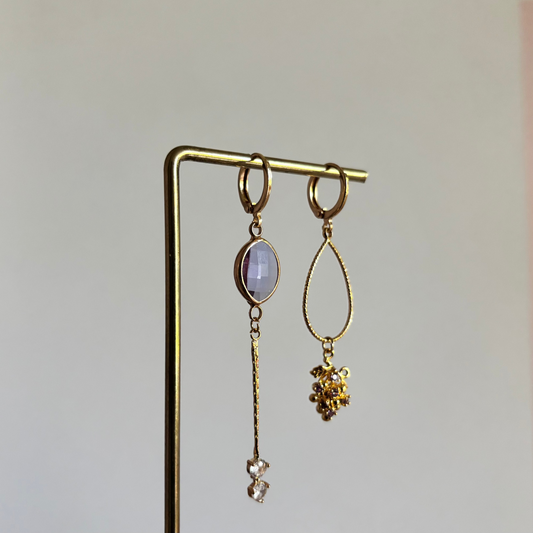 Set asymmetrische goudkleurige oorbellen aan een display. Links een ronde lila facetgeslepen steen met daaronder een fijn goudkleurig staafje en twee transparante steentjes. Rechts een grote opengewerkte druppelvorm met onderaan een bedel in de vorm van een druiventros.