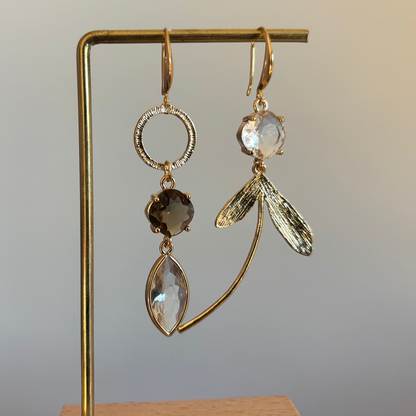 "Asymmetrische 14k vergulde oorbellen met rookbruine en heldere kristallen hangers en goudkleurige blaadjes op een gouden standaard."