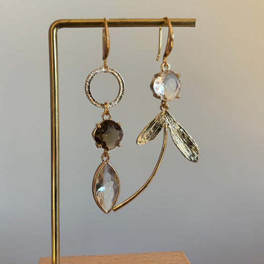 "Asymmetrische 14k vergulde oorbellen met rookbruine en heldere kristallen hangers en goudkleurige blaadjes op een gouden standaard."