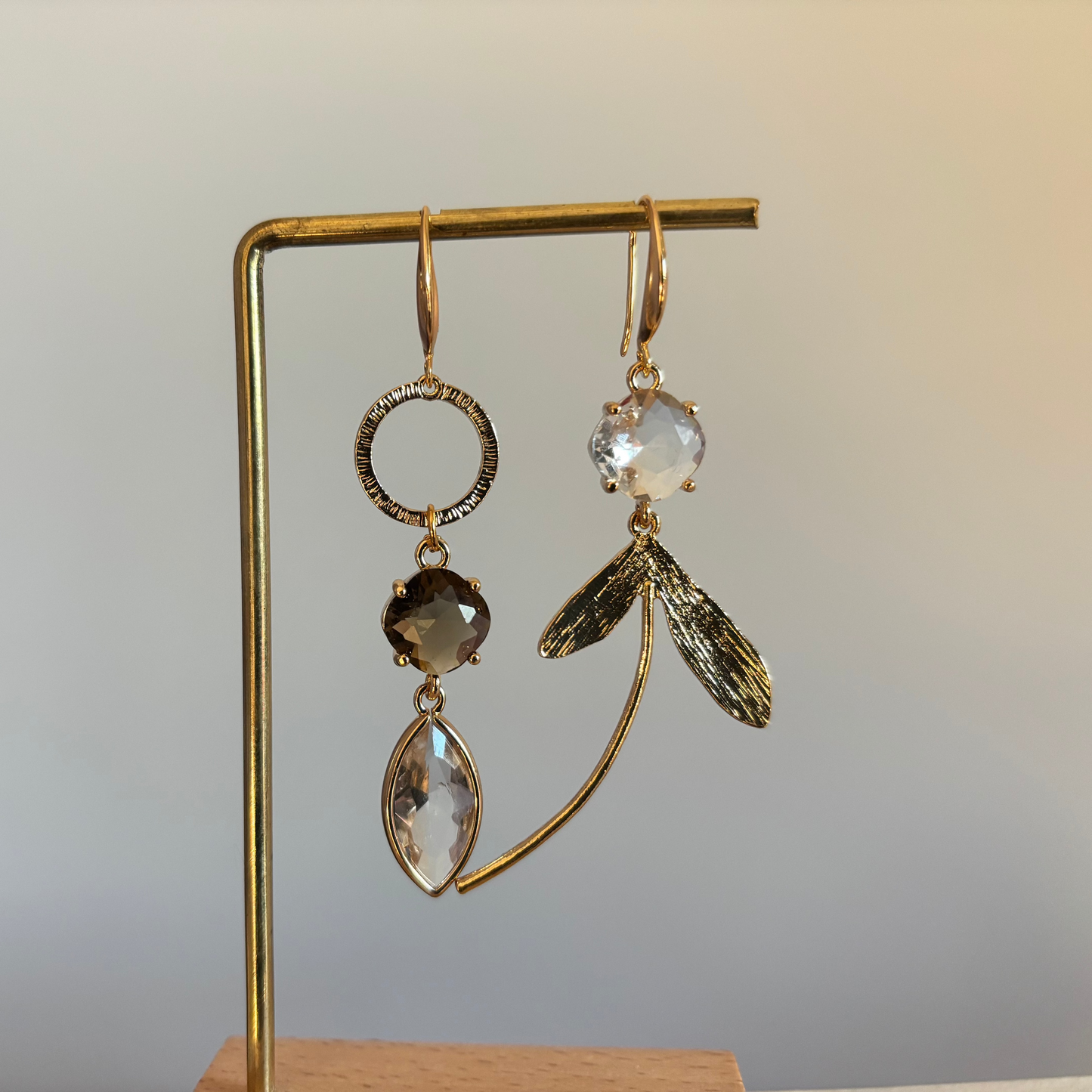 "Asymmetrische 14k vergulde oorbellen met rookbruine en heldere kristallen hangers en goudkleurige blaadjes op een gouden standaard."