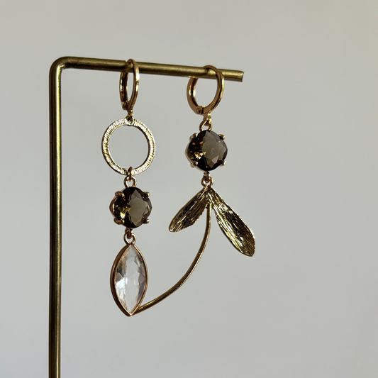 "Asymmetrische 14k vergulde oorbellen met rookbruine en heldere kristallen hangers en goudkleurige blaadjes op een gouden standaard."