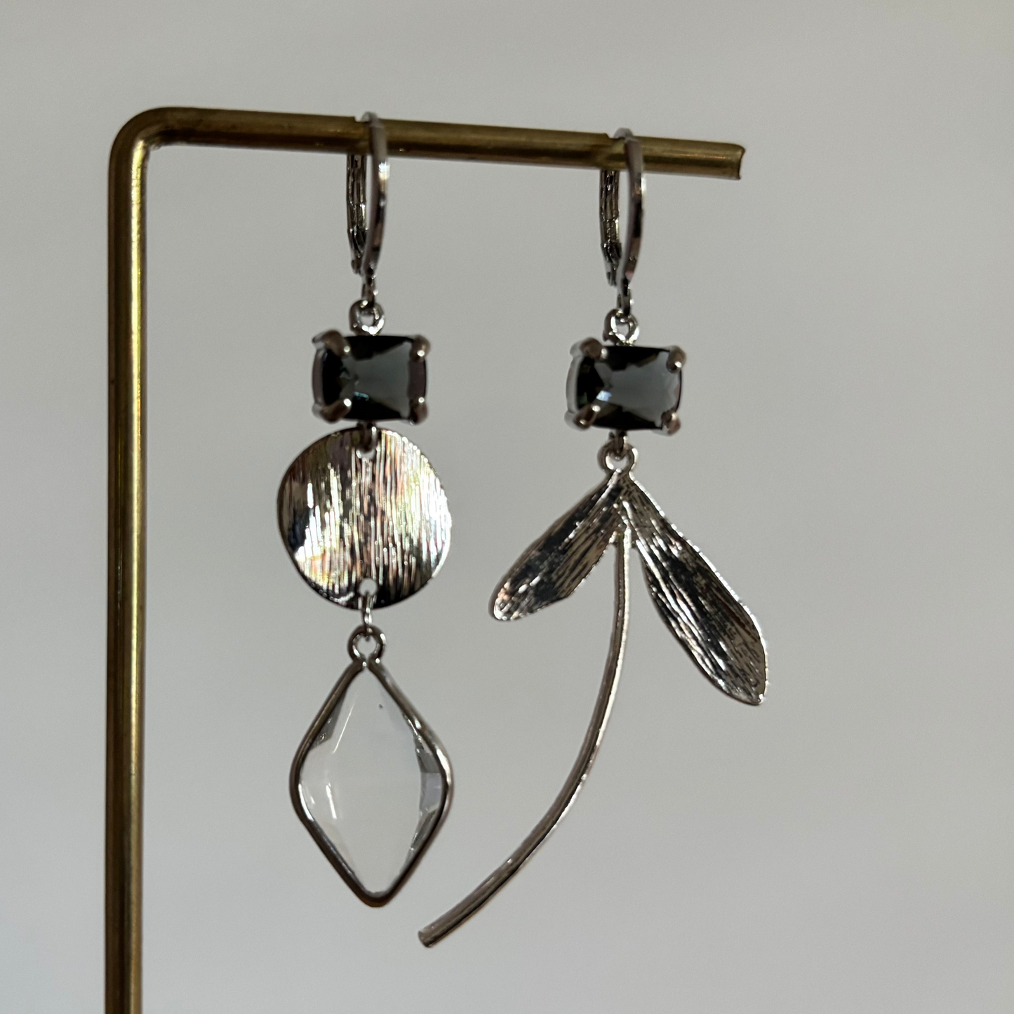 Asymmetrische zilverkleurige oorbellen Cleo aan een rekje. Beide oorbellen hebben bovenaan een rechthoekige donkergrijze kristalsteen; links hangt een gehamerd muntje met een ruitvormig helder kristal, rechts een botanische bladhanger.