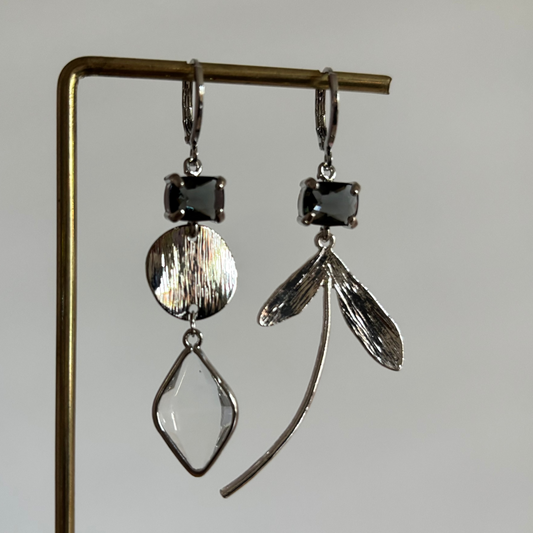 Asymmetrische zilverkleurige oorbellen Cleo aan een rekje. Beide oorbellen hebben bovenaan een rechthoekige donkergrijze kristalsteen; links hangt een gehamerd muntje met een ruitvormig helder kristal, rechts een botanische bladhanger.