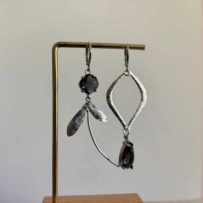 Asymmetrische zilverkleurige oorbellen met rookgrijze stenen, bestaande uit een ruitvormige hanger en een zijde met blaadjes en een lange gebogen metalen lijn.
