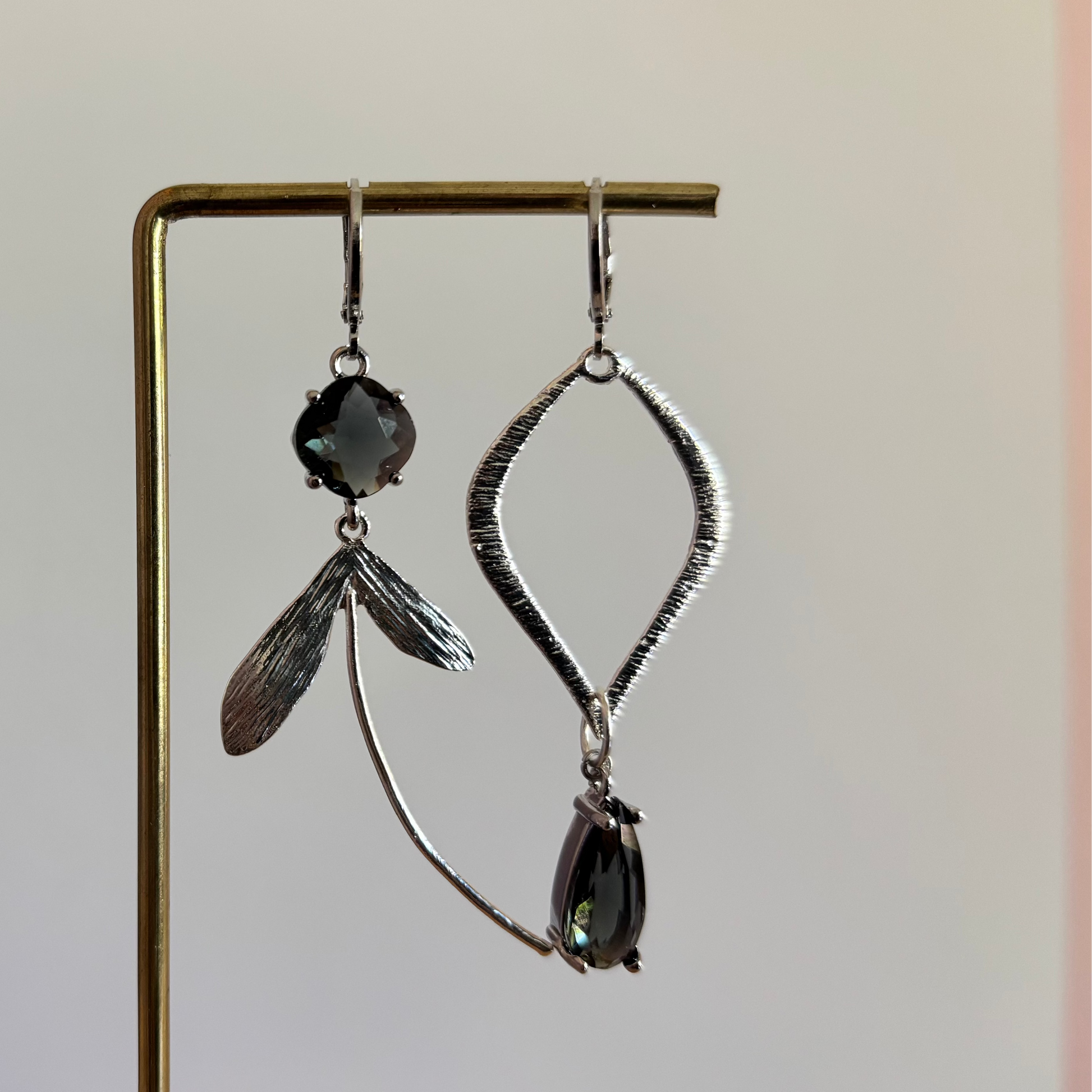 Asymmetrische zilverkleurige oorbellen met rookgrijze stenen, bestaande uit een ruitvormige hanger en een zijde met blaadjes en een lange gebogen metalen lijn.