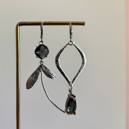 Asymmetrische zilverkleurige oorbellen met rookgrijze stenen, bestaande uit een ruitvormige hanger en een zijde met blaadjes en een lange gebogen metalen lijn.