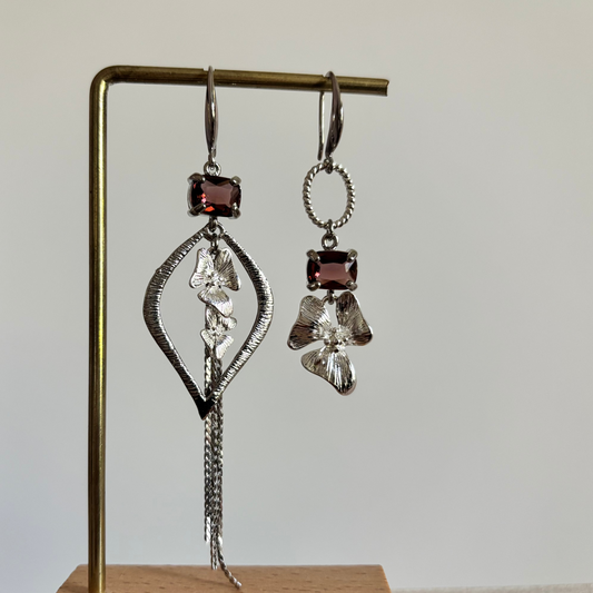 Zilverkleurige asymmetrische hangoorbellen Dani Zilver met rechthoekige paarsrode kristallen, bewerkte ruit met kettinkjes en een zilveren bloemhanger.