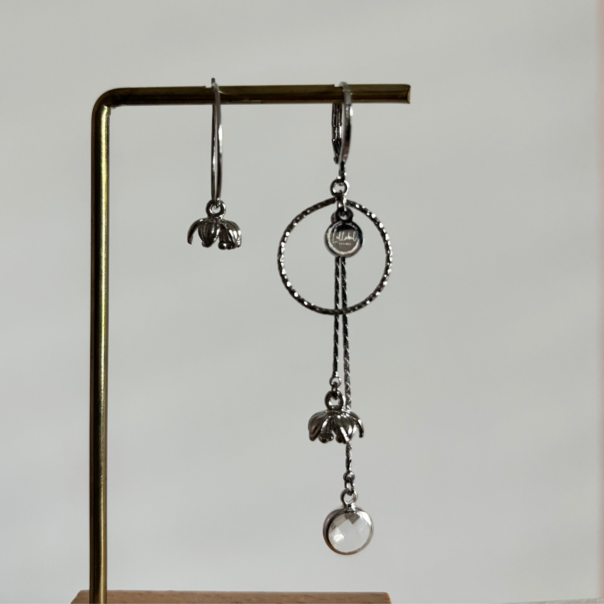Asymmetrische zilverkleurige oorbellen Daphne mismatch aan een rekje. Links een korte hanger met een klein tulpvormig bloemetje; rechts een grote bewerkte cirkel met lange kettinkjes, een bloemetje en een ronde heldere kristalsteen.