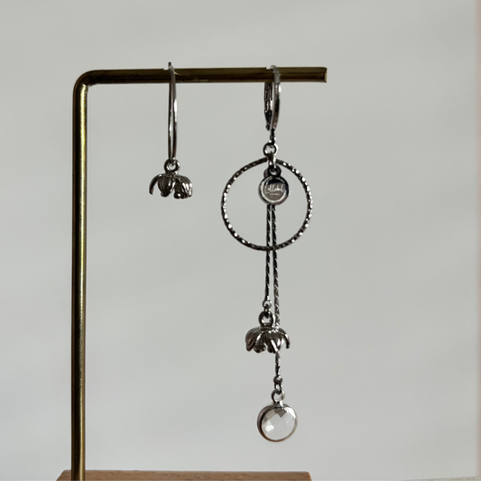 Asymmetrische zilverkleurige oorbellen Daphne mismatch aan een rekje. Links een korte hanger met een klein tulpvormig bloemetje; rechts een grote bewerkte cirkel met lange kettinkjes, een bloemetje en een ronde heldere kristalsteen.