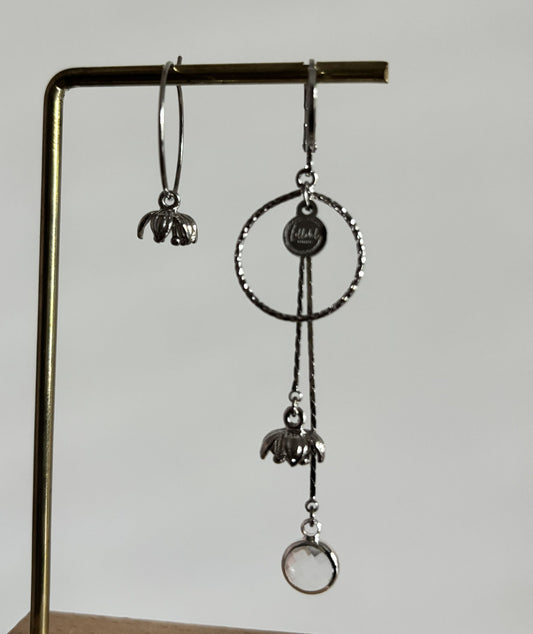 Asymmetrische zilverkleurige oorbellen Daphne mismatch aan een rekje. Links een korte hanger met een klein tulpvormig bloemetje; rechts een grote bewerkte cirkel met lange kettinkjes, een bloemetje en een ronde heldere kristalsteen.