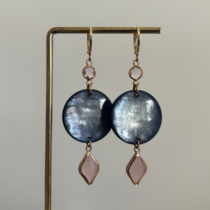 Lange gouden oorbellen bestaande uit een kleine ronde roze steen, een grote ronde donkerblauwe disc met parelmoerglans en onderaan een ruitvormige hanger in melkachtig roze.