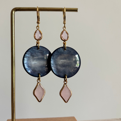 Lange gouden oorbellen bestaande uit een kleine ronde roze steen, een grote ronde donkerblauwe disc met parelmoerglans en onderaan een ruitvormige hanger in melkroze