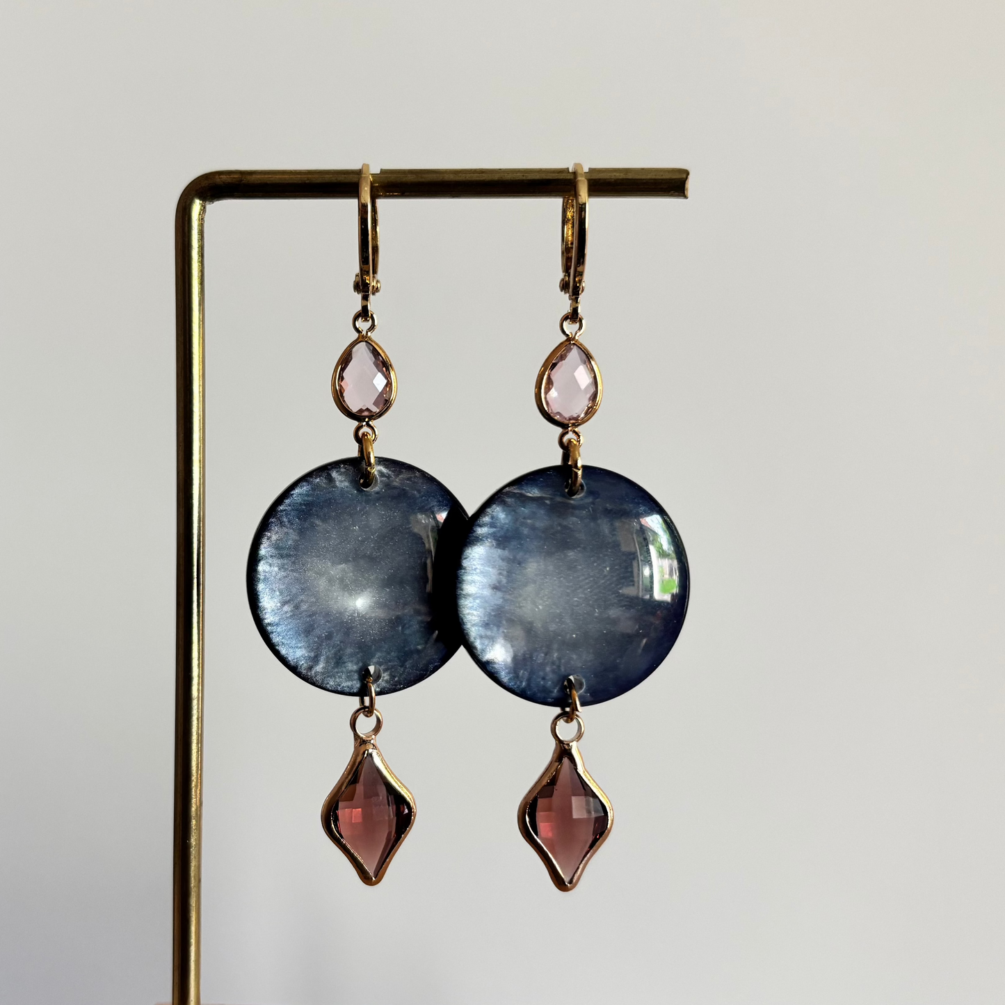 Lange gouden oorbellen bestaande uit een kleine ronde roze steen, een grote ronde donkerblauwe disc met parelmoerglans en onderaan een ruitvormige hanger in burgundy