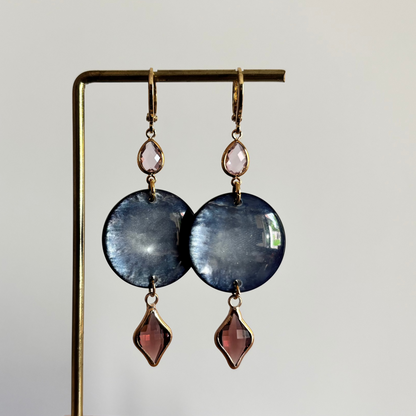 Lange gouden oorbellen bestaande uit een kleine ronde roze steen, een grote ronde donkerblauwe disc met parelmoerglans en onderaan een ruitvormige hanger in burgundy