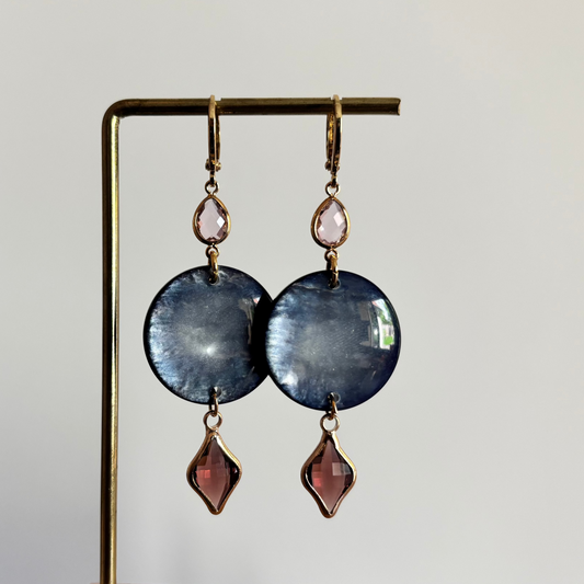 Lange gouden oorbellen bestaande uit een kleine ronde roze steen, een grote ronde donkerblauwe disc met parelmoerglans en onderaan een ruitvormige hanger in burgundy