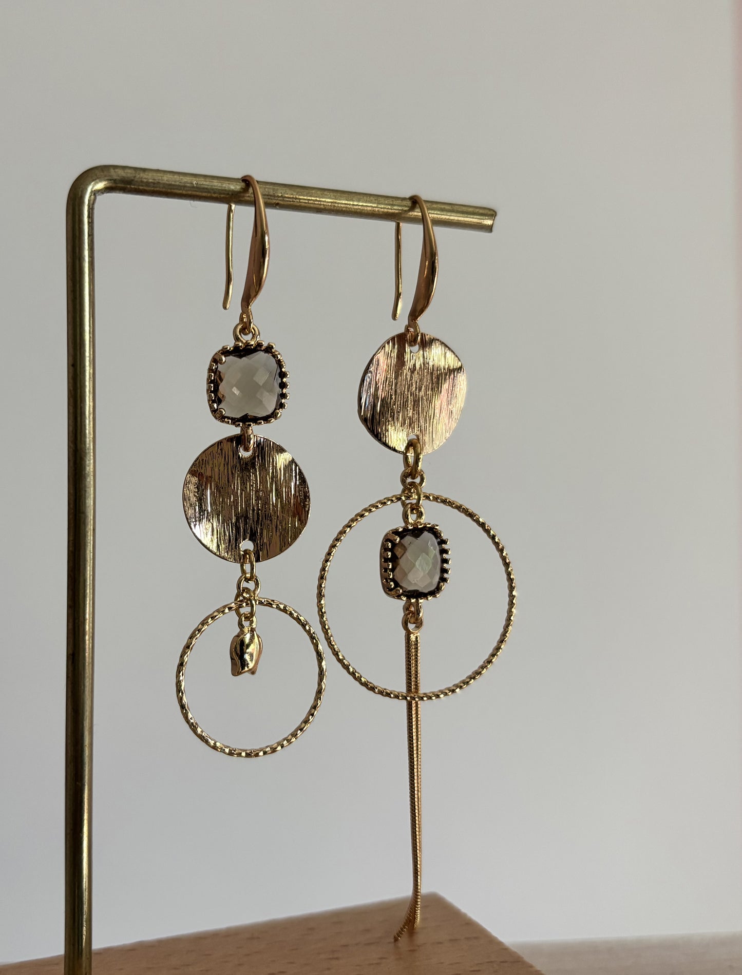 Goudkleurige asymmetrische oorbellen Dewi met ronde geborstelde schijf, bewerkte ring, vierkante taupekleurige kristalsteen en lang kettinkje.