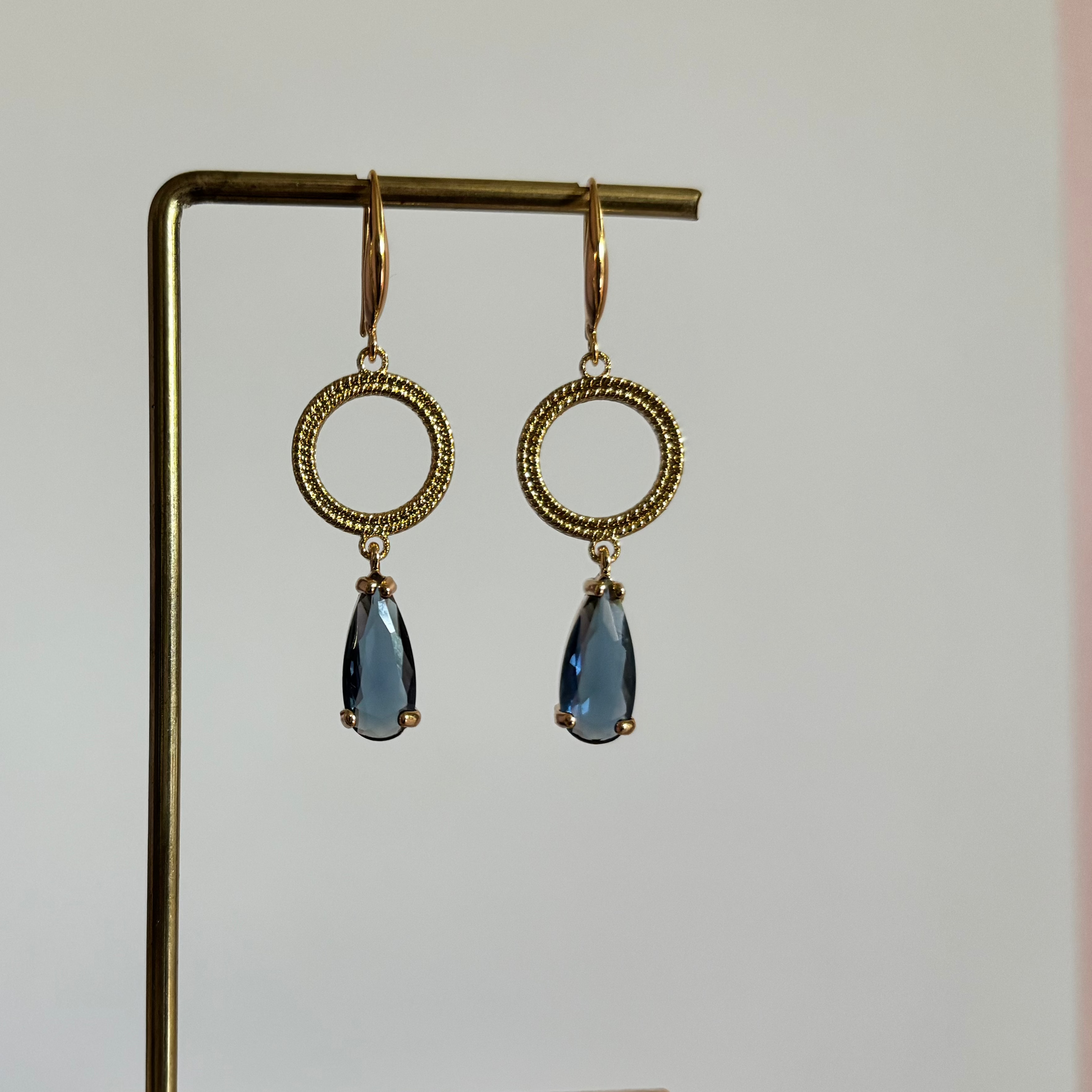 Symmetrische goudkleurige oorbellen Druppel Mir Blauw aan een rekje, met een gedetailleerde, ronde bewerkte ring en een hangende, facetgeslepen donkerblauwe kristaldruppel.