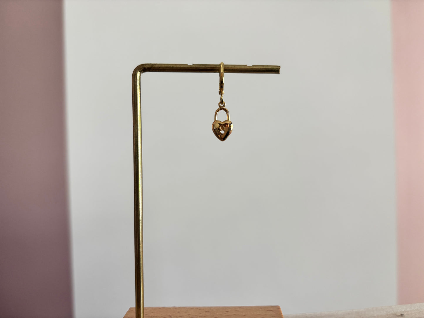 Een goudkleurig klapoorringetje met een hangend hartvormig slotje als bedel, gepresenteerd op een gouden displaystandaard tegen een wit-roze achtergrond.