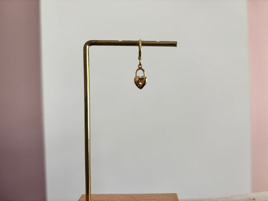 Een goudkleurig klapoorringetje met een hangend hartvormig slotje als bedel, gepresenteerd op een gouden displaystandaard tegen een wit-roze achtergrond.