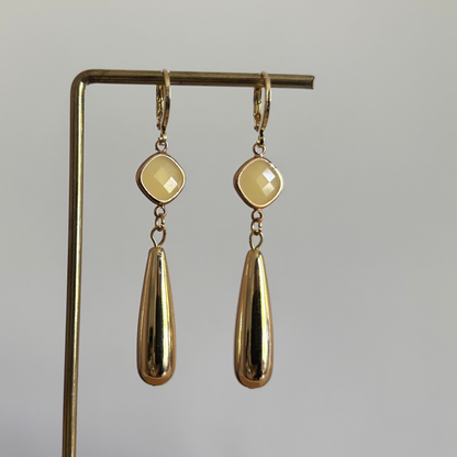 Gouden hanger oorbellen Ellen met matte gele jade-stenen en glanzende goudkleurige druppelhangers op een standaard.