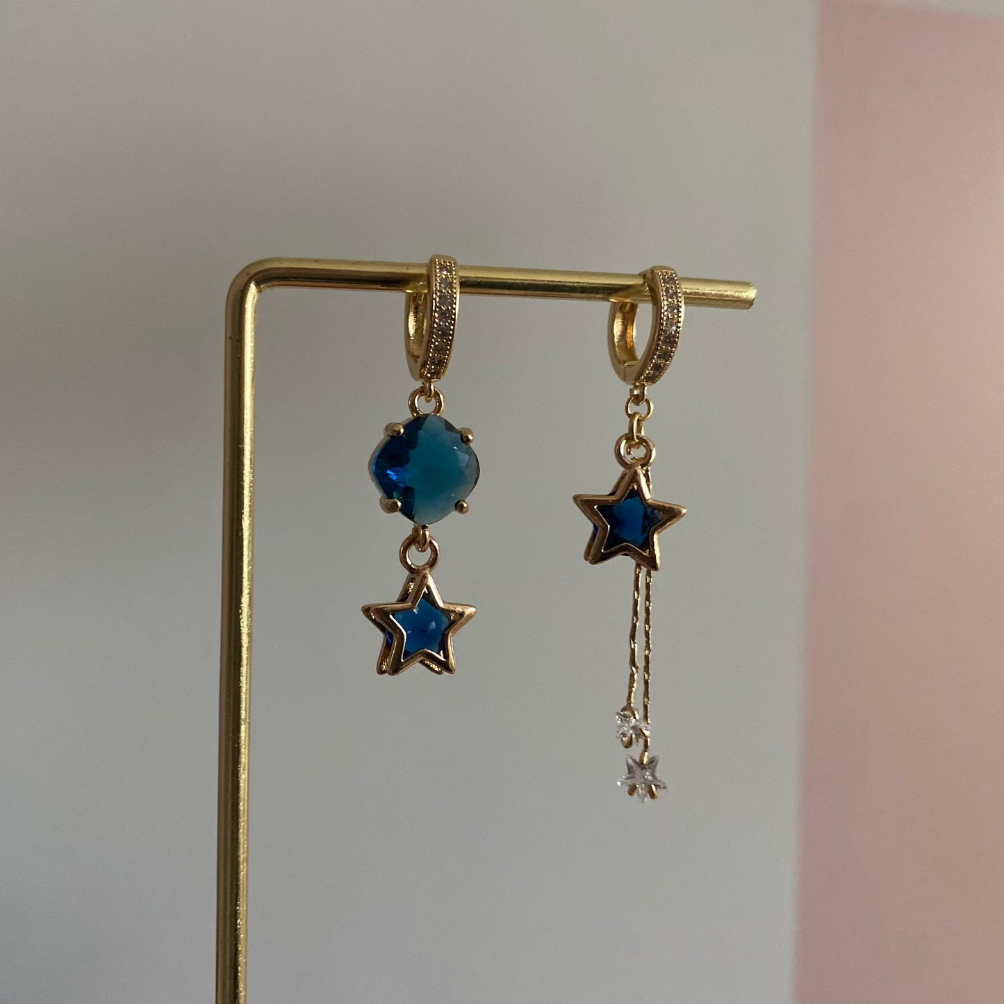 Een foto van het paar oorbellen "Emmeline". Een Asymmetrisch feestelijk oorbellensetje met blauwe glasbedel links en blauwe ster met zirkonia kwastje rechts, beide aan 14k vergulde zirkonia creolen.