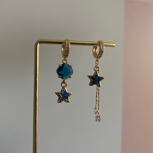 Een foto van het paar oorbellen "Emmeline". Een Asymmetrisch feestelijk oorbellensetje met blauwe glasbedel links en blauwe ster met zirkonia kwastje rechts, beide aan 14k vergulde zirkonia creolen.