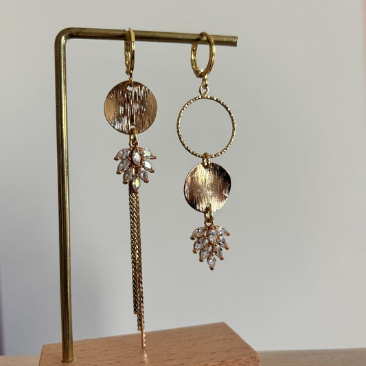 Gouden asymmetrische oorbellen Fabienne met blanke steentjes op een standaard.