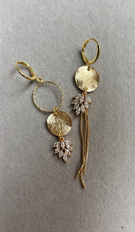 Gouden asymmetrische oorbellen Fabienne met blanke steentjes op een standaard.