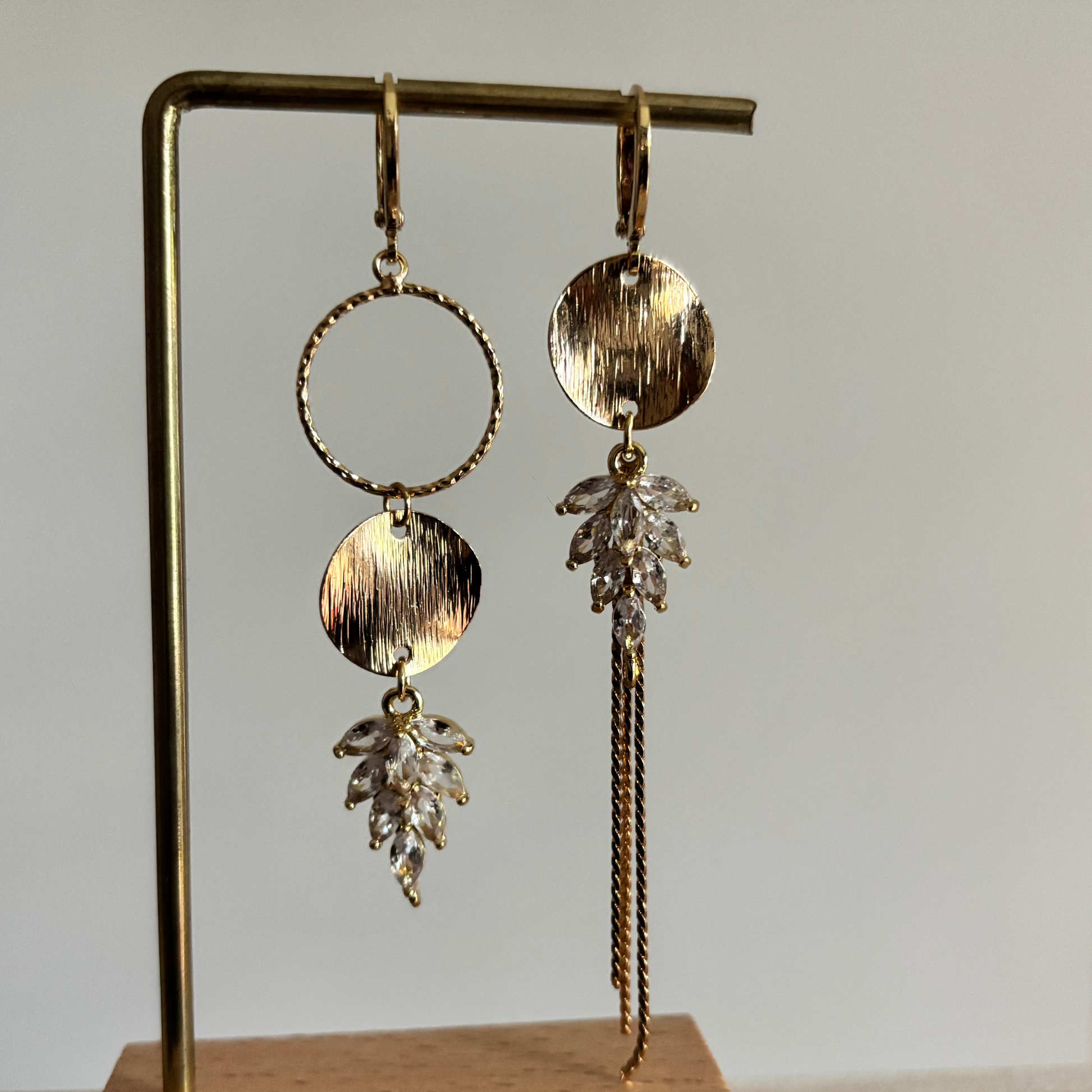 Gouden asymmetrische oorbellen Fabienne met blanke steentjes op een standaard.