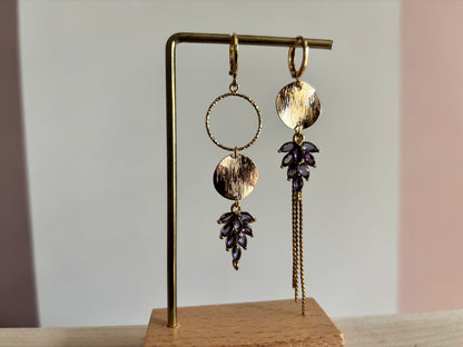 Gouden asymmetrische oorbellen Fabienne met paarse steentjes op een standaard.