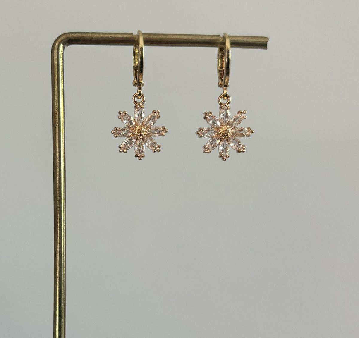 Paar goudkleurige oorbellen Fiet op een standaard, bestaand uit een klapoorringetje met een bloemvormige hanger gemaakt van heldere glinsterende kristalletjes rondom een goudkleurig hart.
