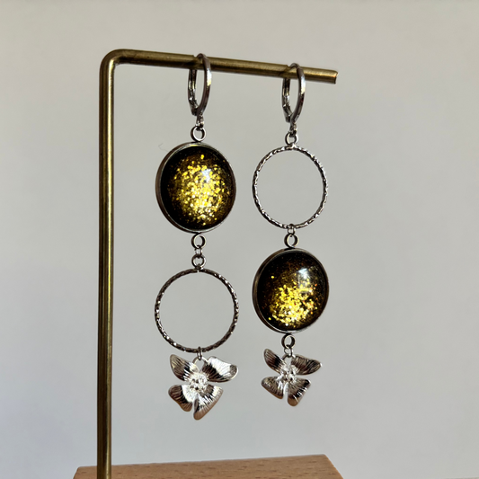 "Zilverkleurige mismatched oorbellen 'Glow Annick Groen Zilver' met ronde olijfgroene glitter cabochons, opengewerkte bewerkte cirkels en zilveren bloembedels aan een sieradenstandaard."