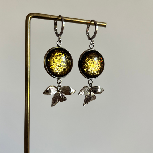 "Symmetrische zilveren oorbellen Glow Blossom met ronde groene glitter cabochons en zilveren bladdetails hangend aan een sieradenstandaard."