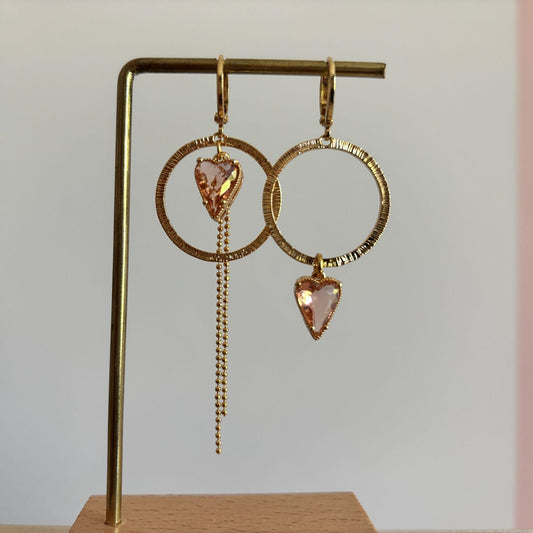 Gouden mismatched oorbellen set Gwen met grote bewerkte ronde cirkels en lichtroze hartvormige glasstenen. Eén oorbel heeft het hartje onderaan de ring, de andere heeft het hartje bovenaan met een lang strass- en bolletjeskettinkje."