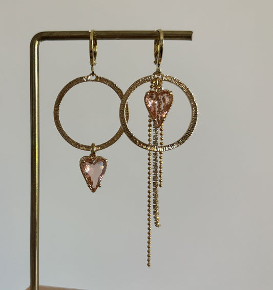 Gouden mismatched oorbellen set Gwen met grote bewerkte ronde cirkels en lichtroze hartvormige glasstenen. Eén oorbel heeft het hartje onderaan de ring, de andere heeft het hartje bovenaan met een lang strass- en bolletjeskettinkje."