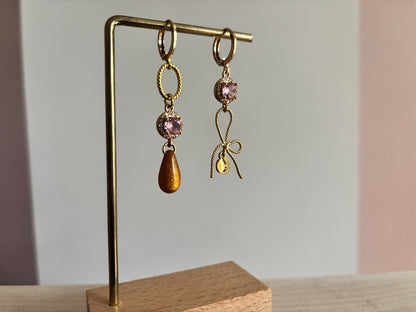 Gouden asymmetrische oorbellen Harper met roze kristal, strikje en okerkleurige druppel op een standaard.