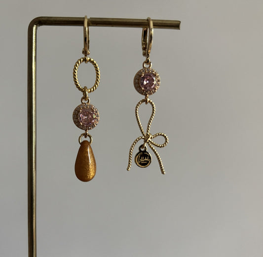 Gouden asymmetrische oorbellen Harper met roze kristal, strikje en okerkleurige druppel op een standaard.
