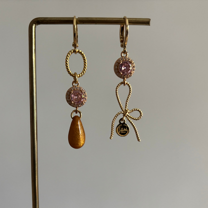 Gouden asymmetrische oorbellen Harper met roze kristal, strikje en okerkleurige druppel op een standaard.