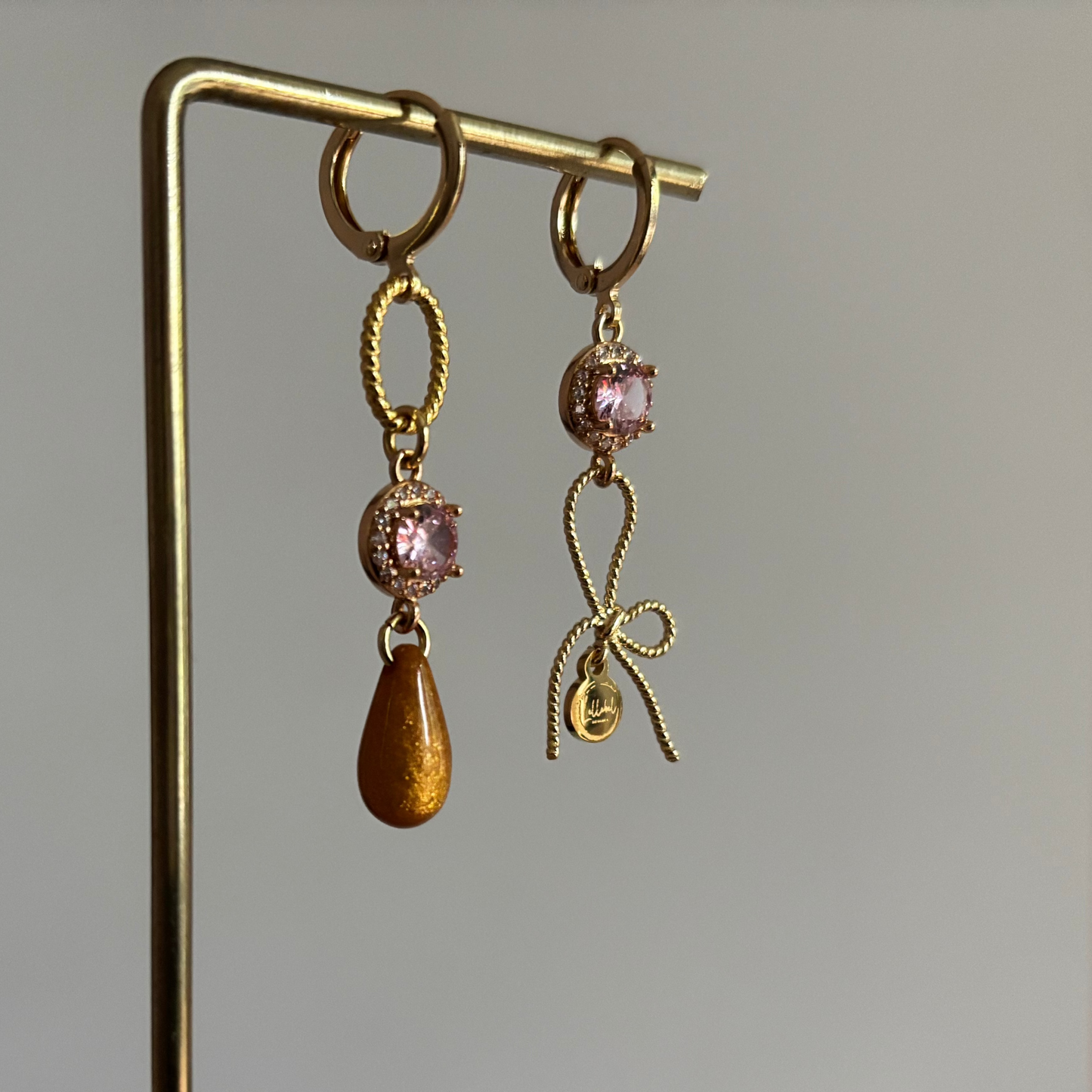 Gouden asymmetrische oorbellen Harper met roze kristal, strikje en okerkleurige druppel op een standaard.