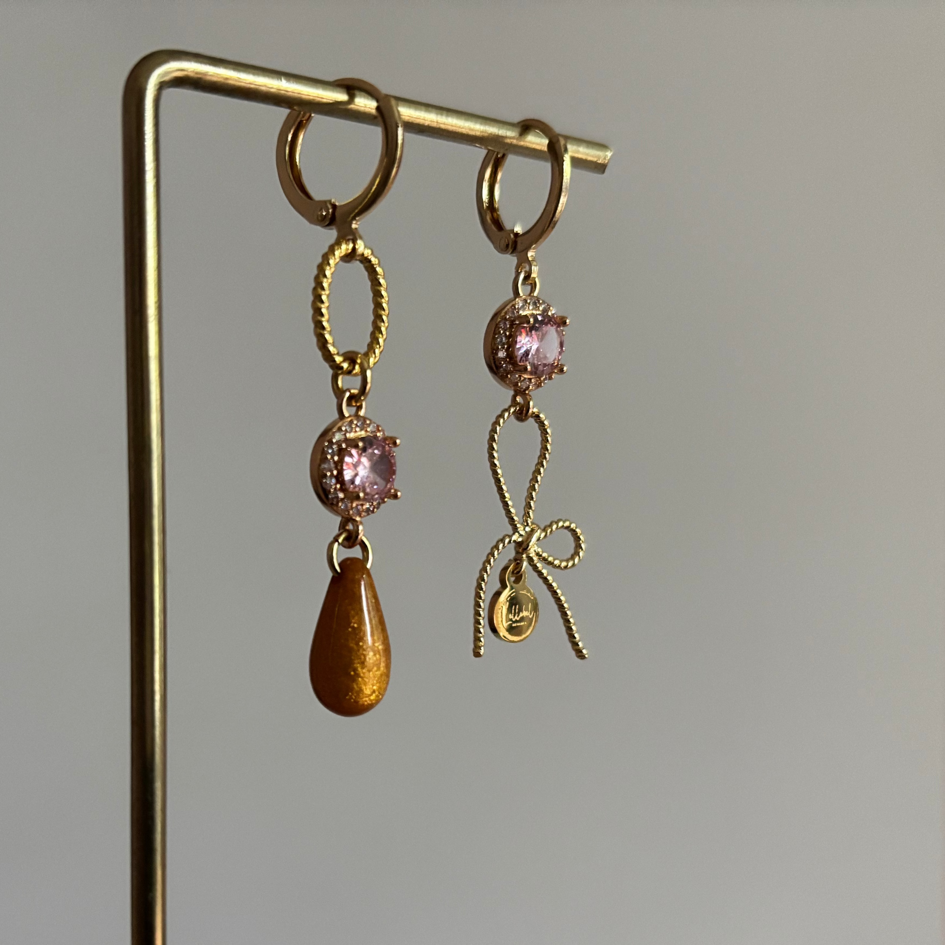 Gouden asymmetrische oorbellen Harper met roze kristal, strikje en okerkleurige druppel op een standaard.