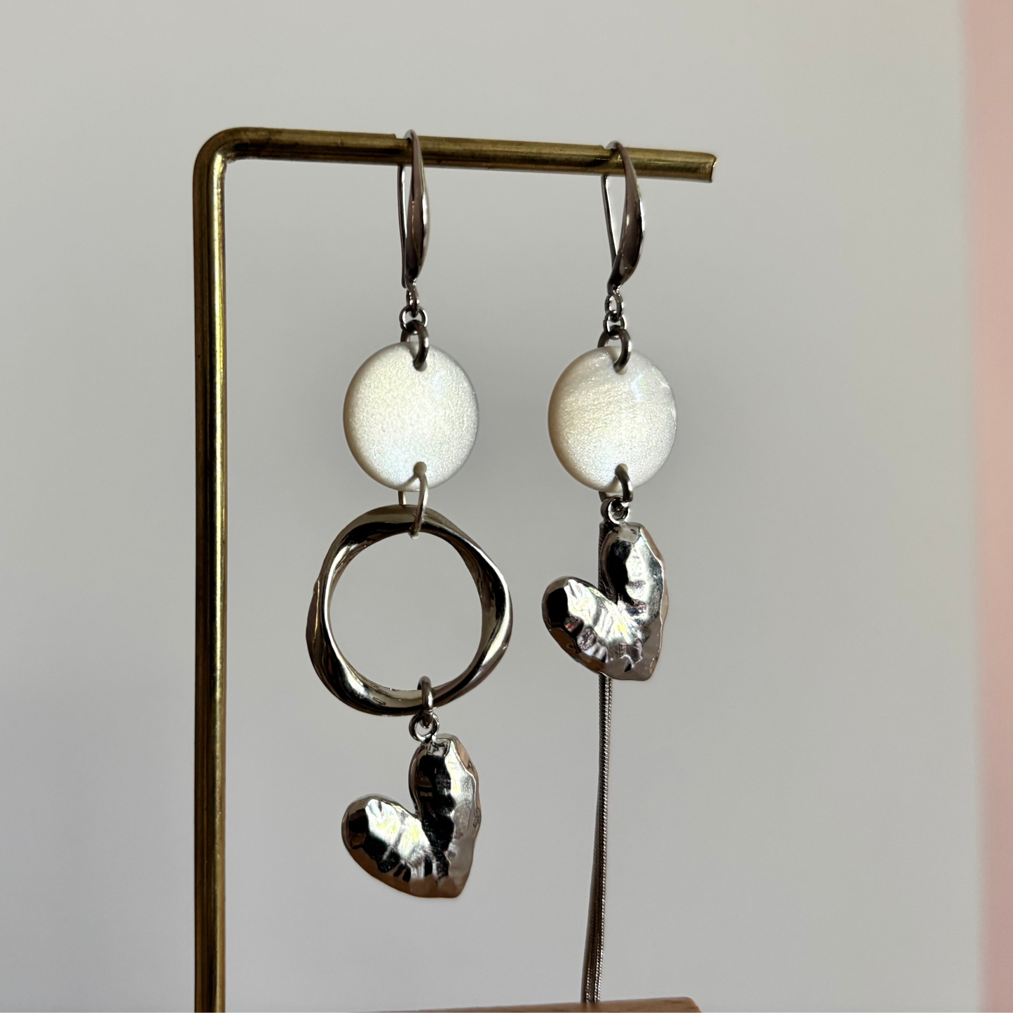 Asymmetrische zilveren oorbellen Ivy met ronde wit parelmoer cabochons, gehamerde ringen en een zilveren hartvormige bedel hangend aan een rekje.