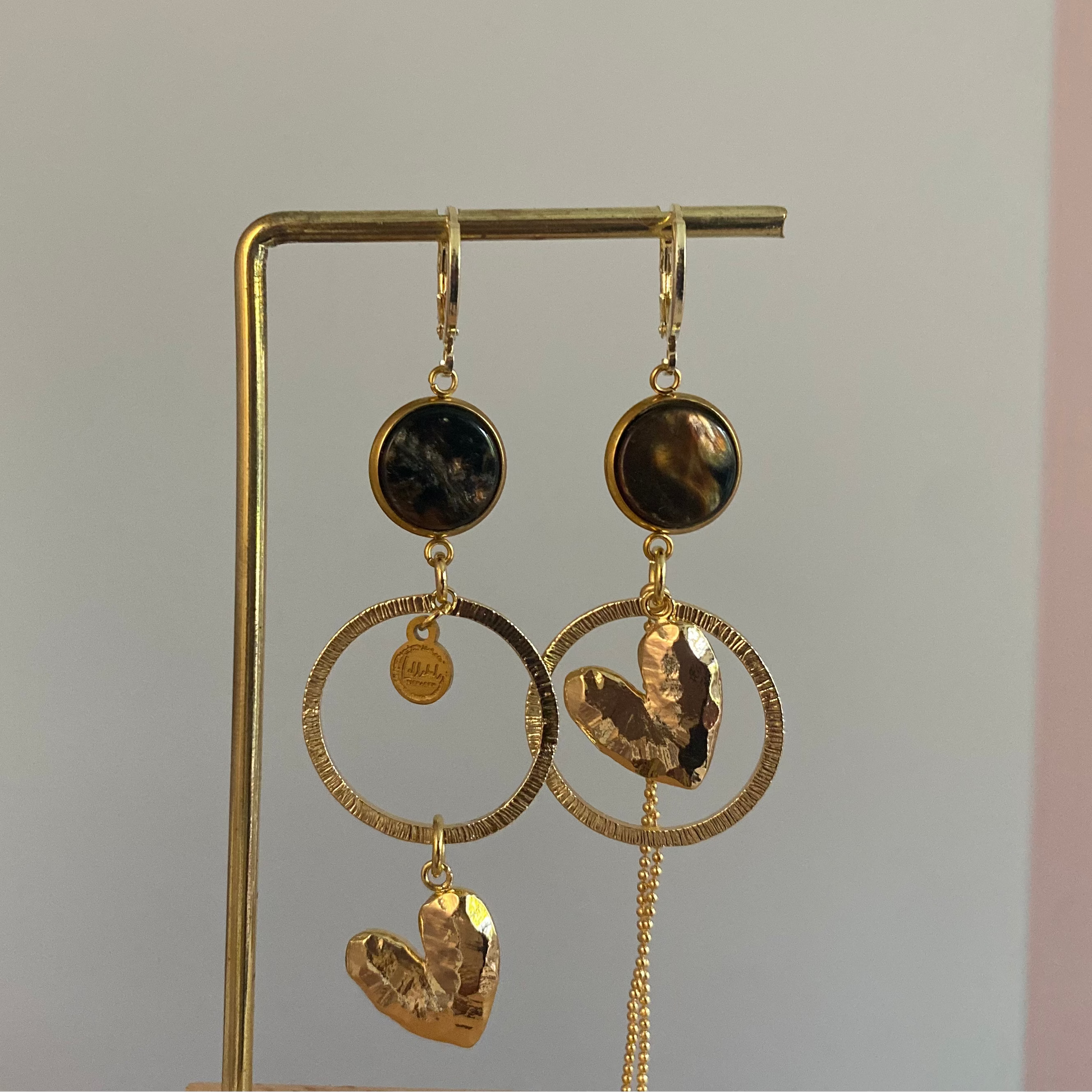 Asymmetrische 14k vergulde oorbellen met donkerbruine cabochon, gehamerde ringen, hartbedel en klein kwastje, ca. 6 cm lang.
