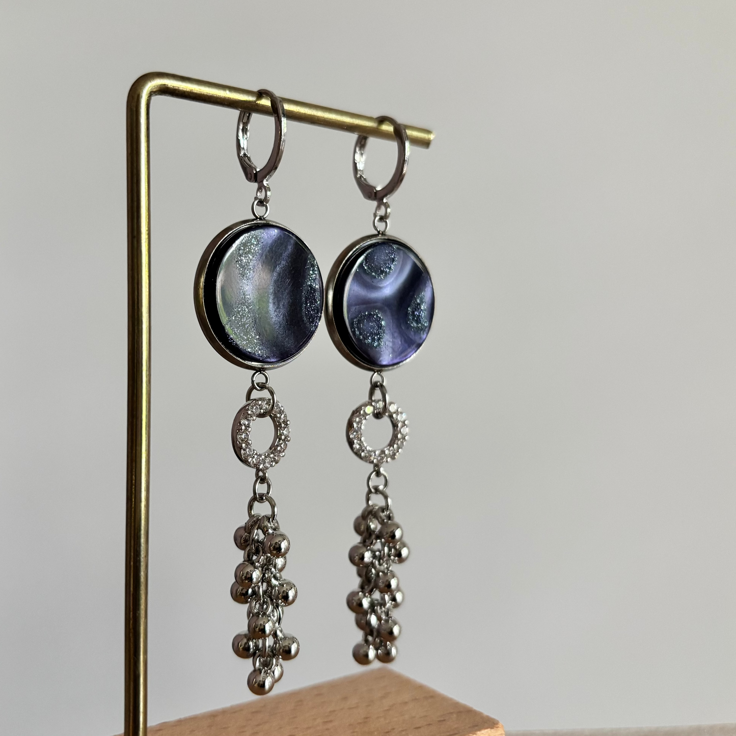Zilverkleurige oorbellen Joke aan een rekje. Model met een ronde donkerblauwe  polaris steen, een klein strass-ringetje en een hangend trosje van zilveren bolletjes.