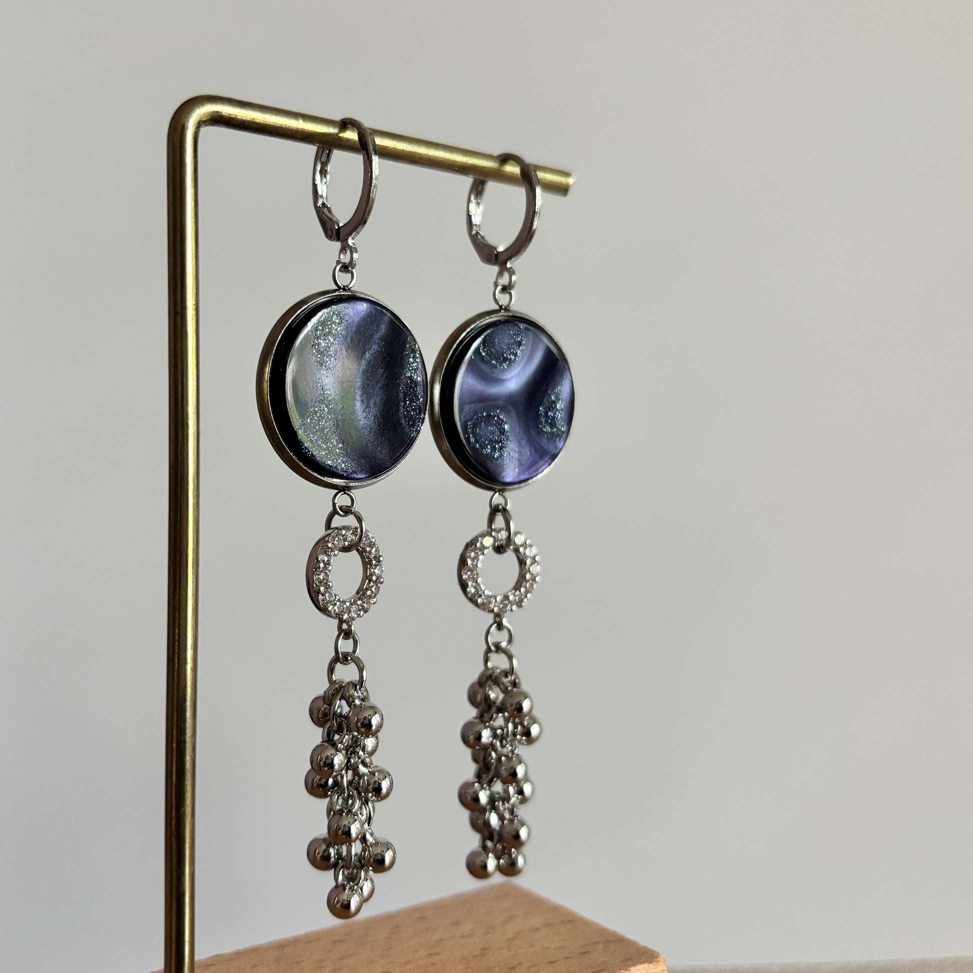 Zilverkleurige oorbellen Joke aan een rekje. Model met een ronde donkerblauwe  polaris steen, een klein strass-ringetje en een hangend trosje van zilveren bolletjes.