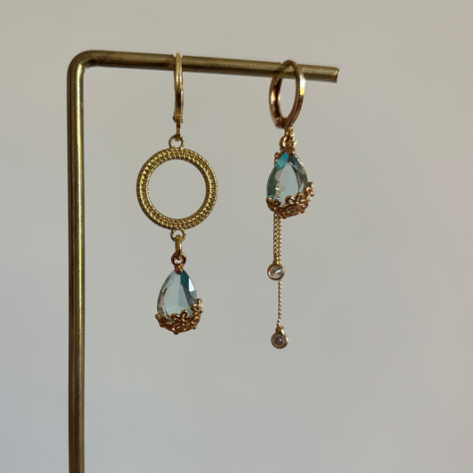 Set asymmetrische goudkleurige oorbellen aan een display. Links een opengewerkte ronde cirkel met daaronder een hangende, lichtblauwe facetgeslepen druppelsteen. Rechts dezelfde blauwe druppelsteen met daaronder twee fijne goudkleurige kettingkjes met kleine steentjes.
