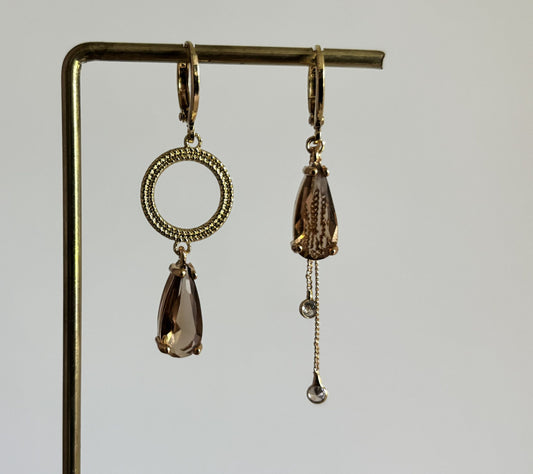 Set asymmetrische goudkleurige oorbellen aan een display. Links een opengewerkte ronde cirkel met daaronder een hangende, rookbruine facetgeslepen druppelsteen. Rechts dezelfde bruine druppelsteen met daaronder twee fijne goudkleurige kettingkjes met kleine steentjes.