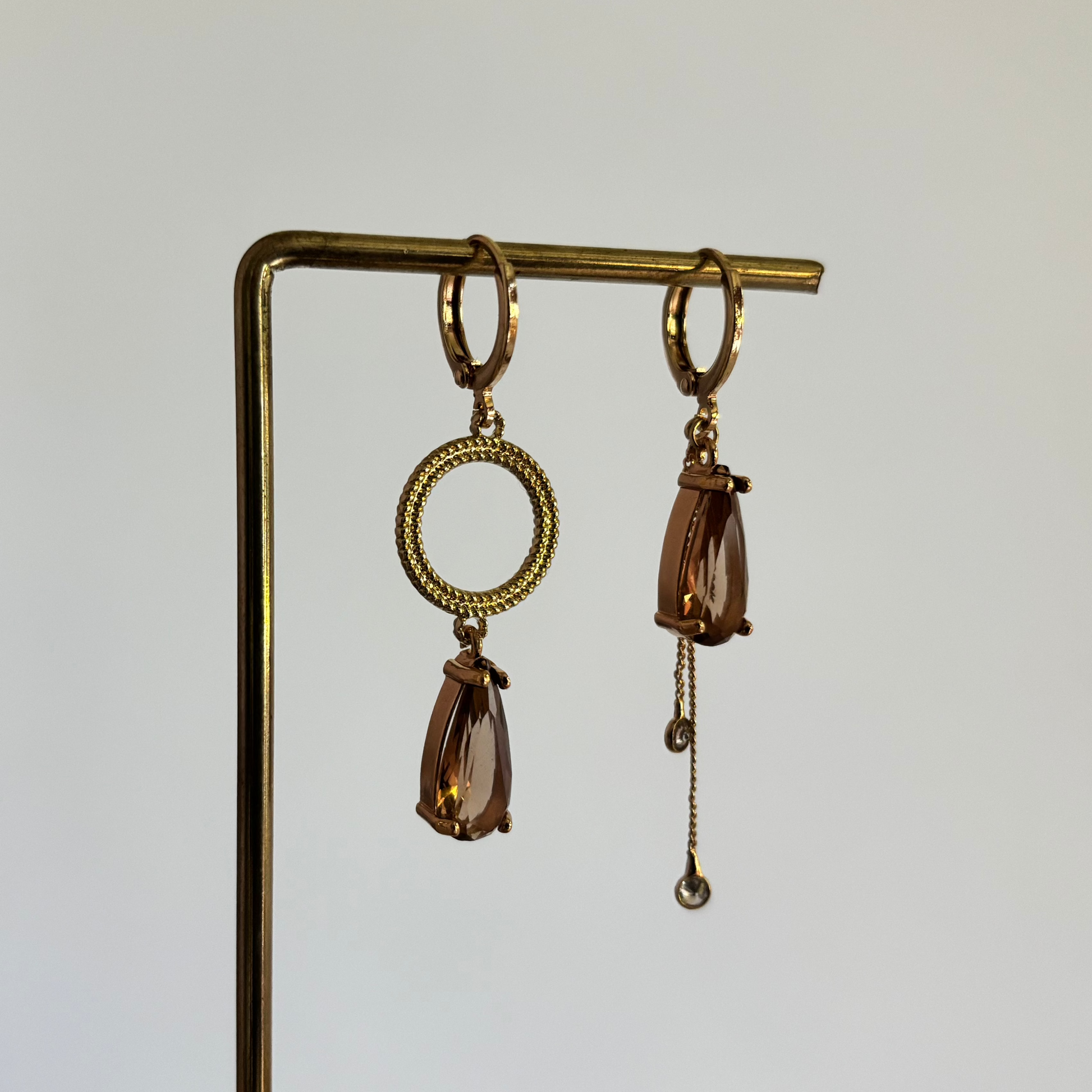 Set asymmetrische goudkleurige oorbellen aan een display. Links een opengewerkte ronde cirkel met daaronder een hangende, rookbruine facetgeslepen druppelsteen. Rechts dezelfde bruine druppelsteen met daaronder twee fijne goudkleurige kettingkjes met kleine steentjes.