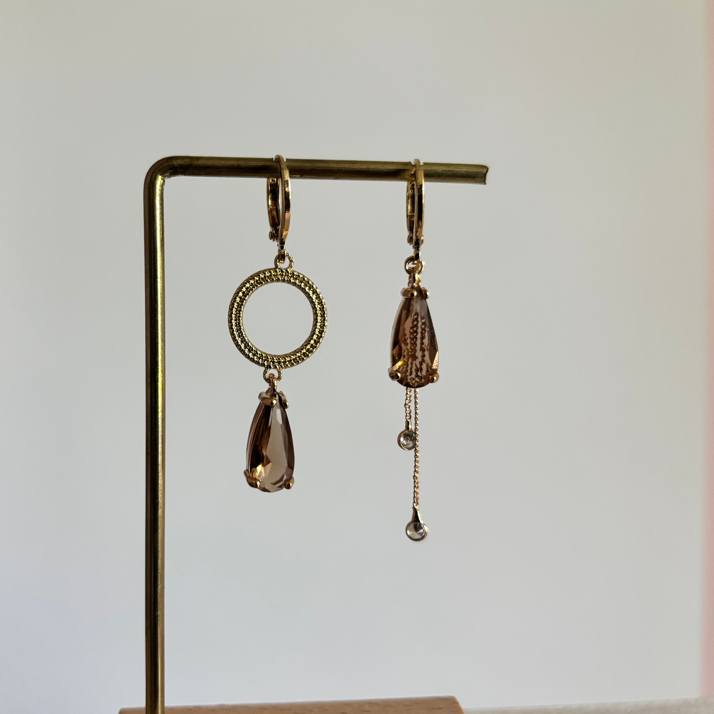 Set asymmetrische goudkleurige oorbellen aan een display. Links een opengewerkte ronde cirkel met daaronder een hangende, rookbruine facetgeslepen druppelsteen. Rechts dezelfde bruine druppelsteen met daaronder twee fijne goudkleurige kettingkjes met kleine steentjes.