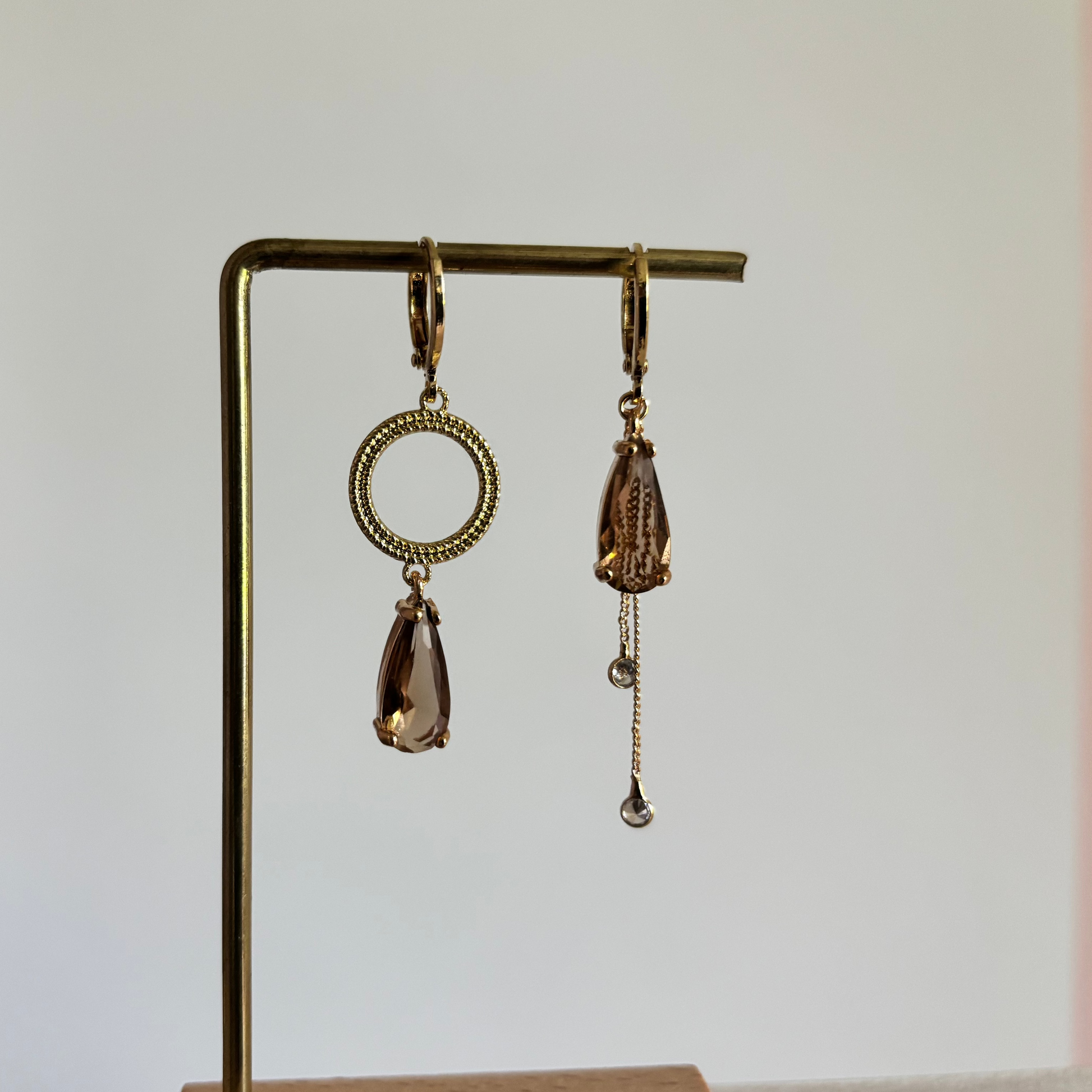 Set asymmetrische goudkleurige oorbellen aan een display. Links een opengewerkte ronde cirkel met daaronder een hangende, rookbruine facetgeslepen druppelsteen. Rechts dezelfde bruine druppelsteen met daaronder twee fijne goudkleurige kettingkjes met kleine steentjes.